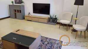 Een moderne woonkamer met een flatscreen televisie, een salontafel, twee witte fauteuils, een dressoir en een klok aan de muur.