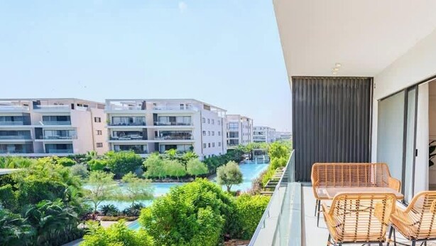 A balcony with wicker furniture overlooking a swimming pool and greenery, with modern buildings in the background. - Luxury 3BR • Lakeview Residence • Premium Stay (القاهرة)