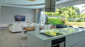 Una cocina moderna con una isla central, zona de comedor con mobiliario blanco y un amplio ventanal con vistas a un jardín.