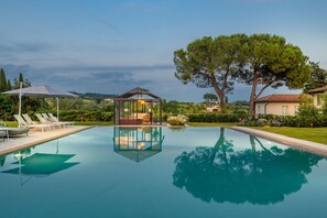 Ein Swimmingpool mit Liegestühlen, einem gläsernen Pavillon und einem großen Baum.