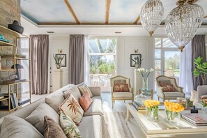 Ein geräumiges Wohnzimmer mit einer großen Couch, einem Couchtisch und zwei Sesseln. Es gibt Bücherregale, eine Chandelier und einen Blick ins Freie.