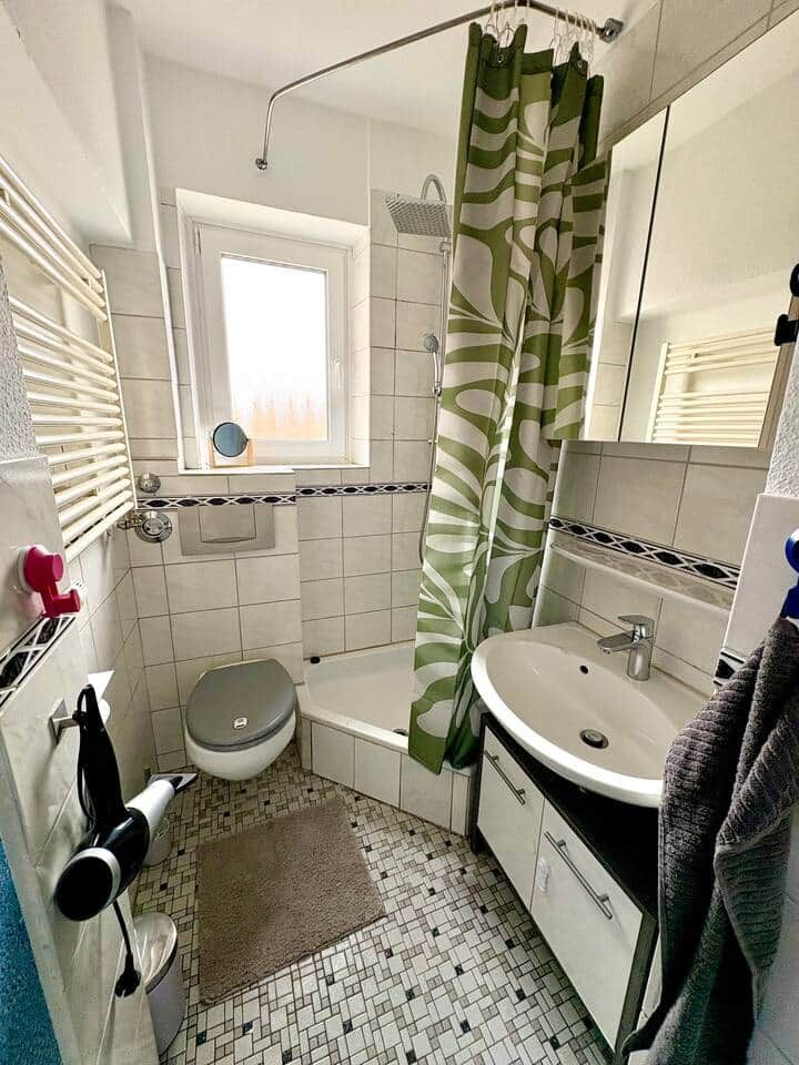 Baño con inodoro, lavamanos y ducha. Hay una ventana sobre el inodoro y un perchero con una toalla.