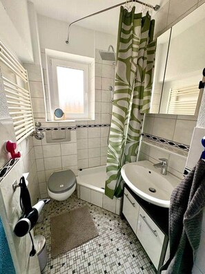 Un bagno con il water, un lavabo e una doccia. Si vede una finestra sopra il water e un portasciugamani con un asciugamano.