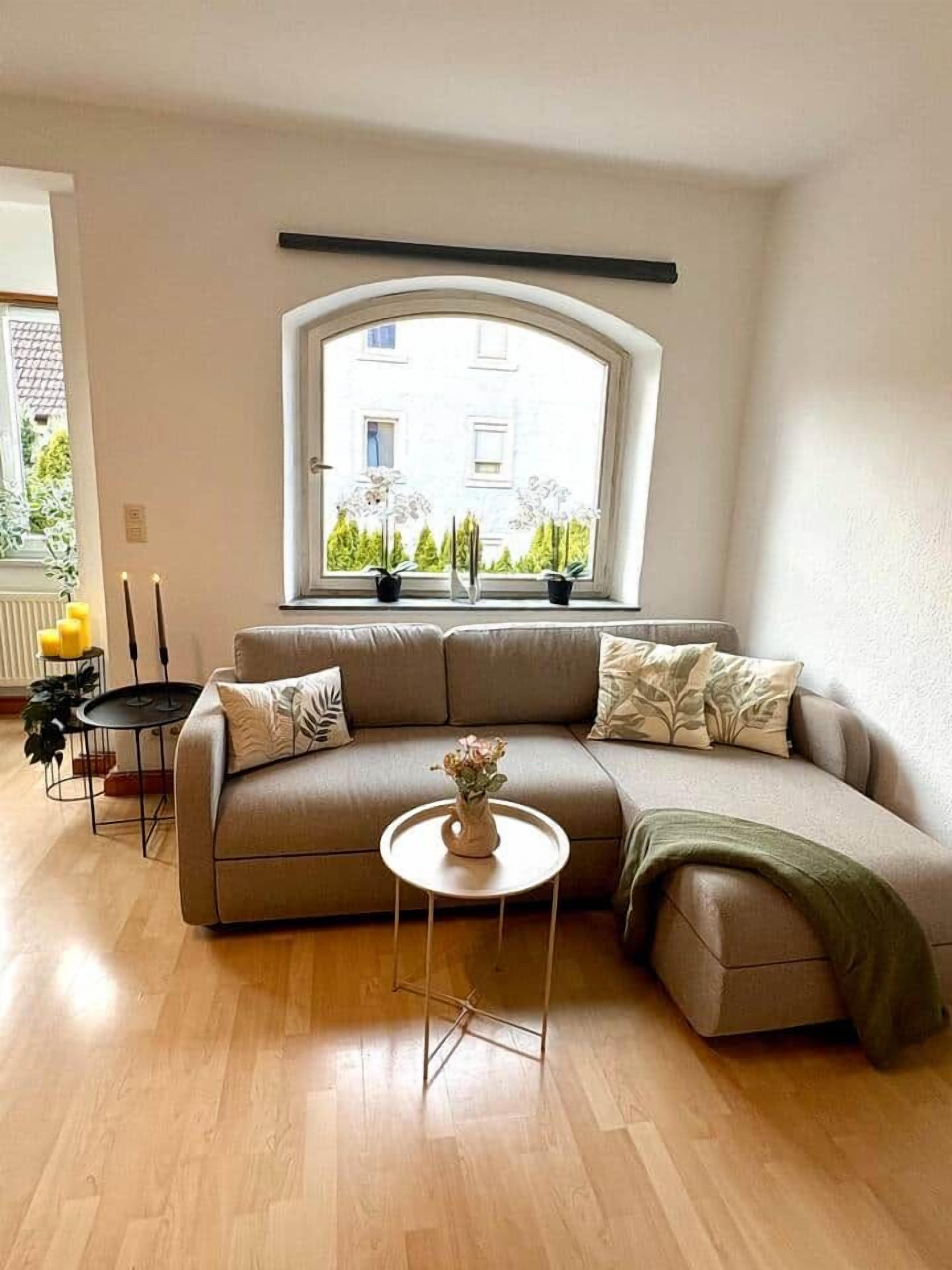 Una sala de estar moderna con un sofá curvo, una mesita redonda y una ventana con vistas a zonas verdes y edificios.