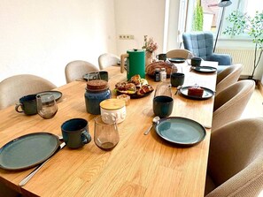 Ein Esstisch ist gedeckt mit Tellern, Tassen und einer Kaffeekanne in einem hellen Raum mit Fenster und Pflanze im Hintergrund.