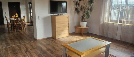 Ein Wohnzimmer mit Holzboden, einem an der Wand befestigten Fernseher, einem Holzschrank, einem Esstisch mit Stühlen und einer Topfpflanze.