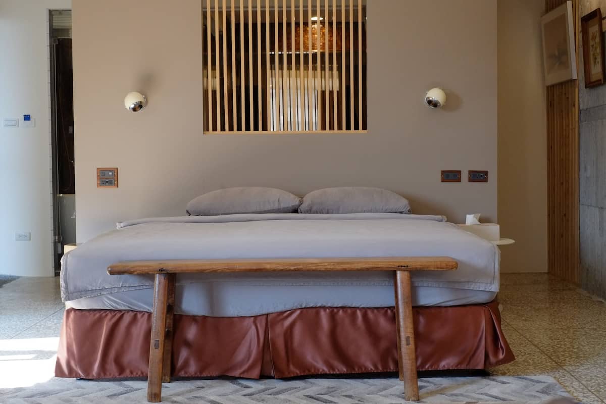 A bedroom with a bed, a wooden bench, a window with blinds, and a wall-mounted light fixture.