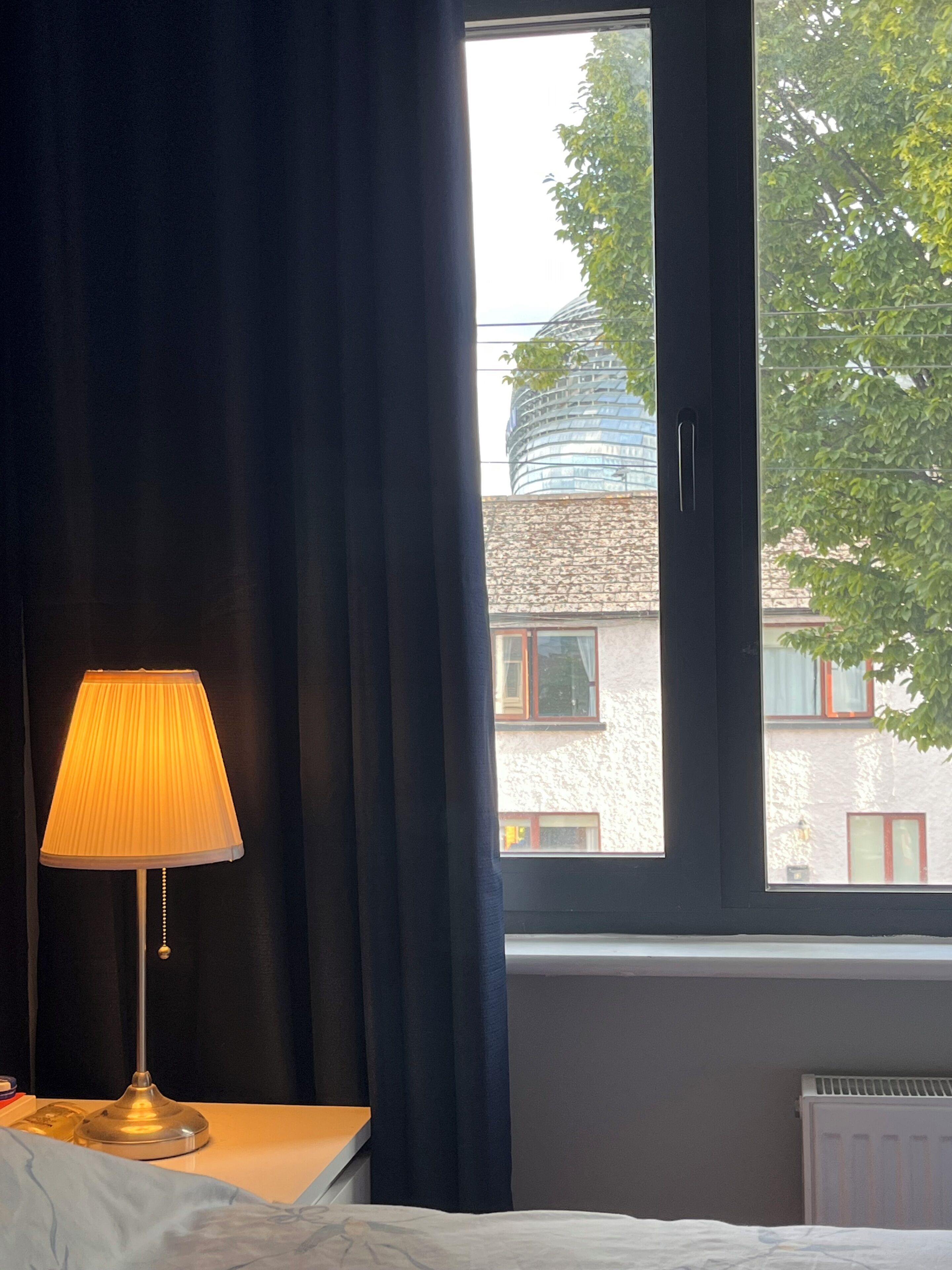 A room with a bed, a bedside table with a lamp, a window with curtains, and a view of a building outside.