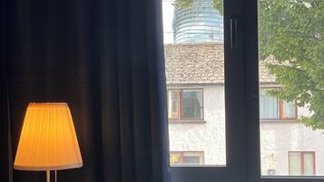 A room with a bed, a bedside table with a lamp, a window with curtains, and a view of a building outside.