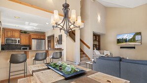 A modern kitchen with stainless steel appliances, a wooden dining table with a centerpiece, and a living area with a gray sofa and a mounted TV. - Elevated Tahoe Escape (Incline Village)