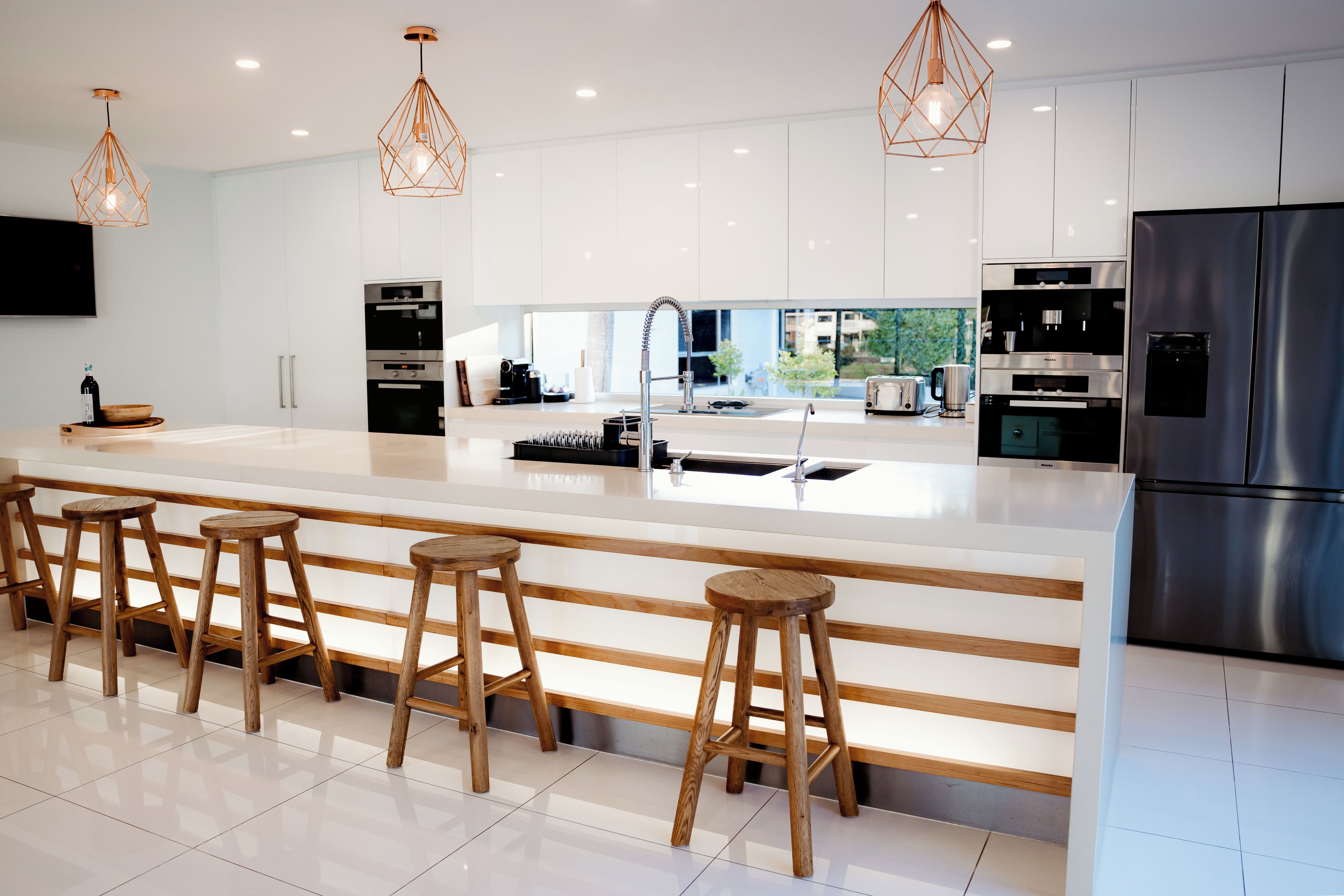 A modern kitchen with a central island, stainless steel appliances, wooden stools, and pendant lighting.