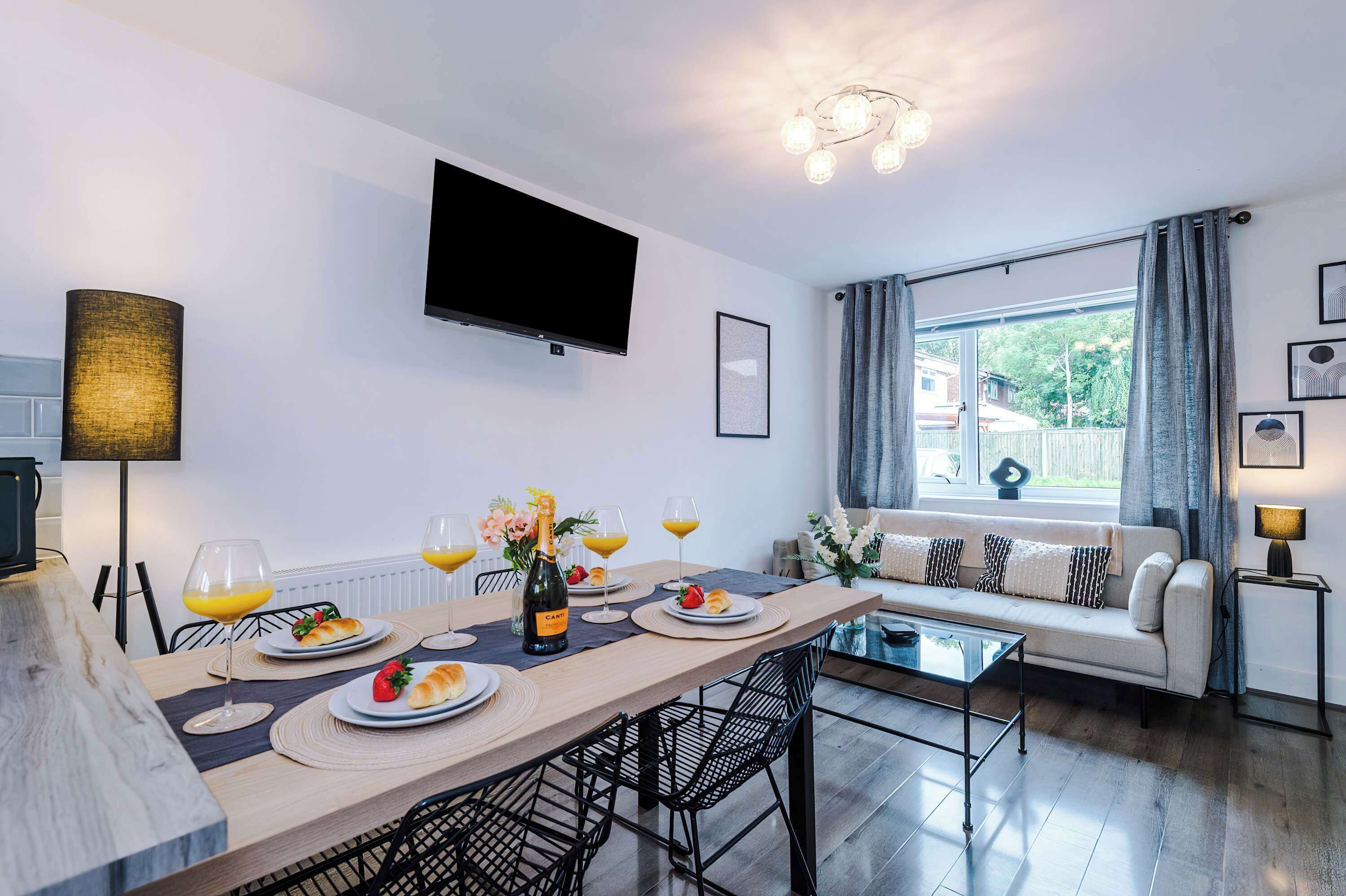 Stylish open-plan living and dining area with modern decor, cozy sofa, wall-mounted TV, and elegant table set for four. Enjoy ambient lighting, framed art, and natural light in a warm, welcoming space perfect for relaxing or entertaining.