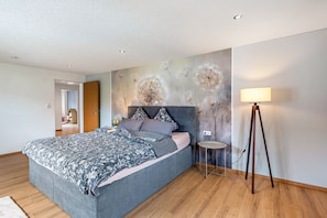 Une chambre moderne avec un grand lit, une table de chevet, une lampe sur pied et un mur orné d’un motif de pissenlit.