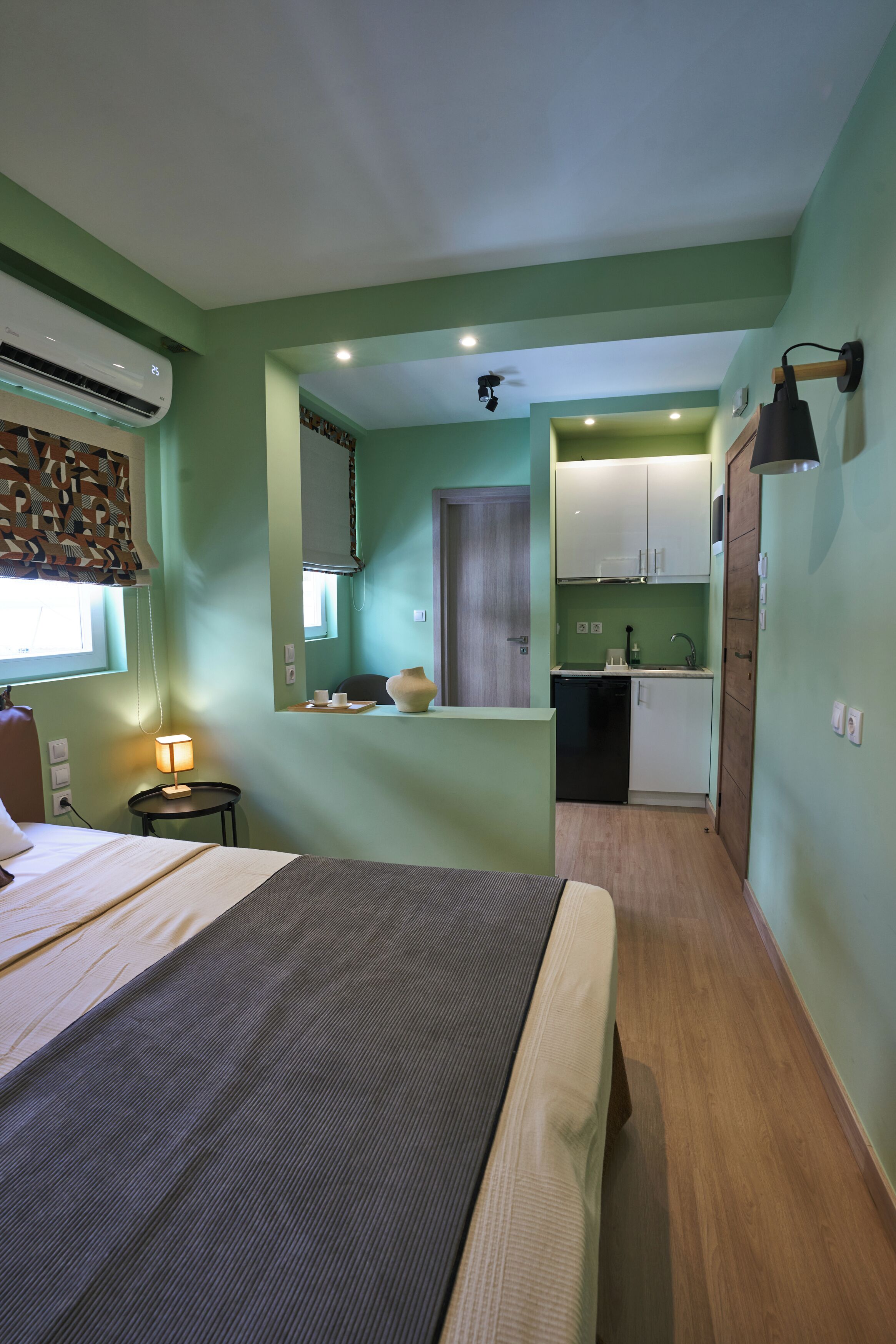 A compact hotel room with a bed, bedside table, lamp, and a view of the kitchen area.
