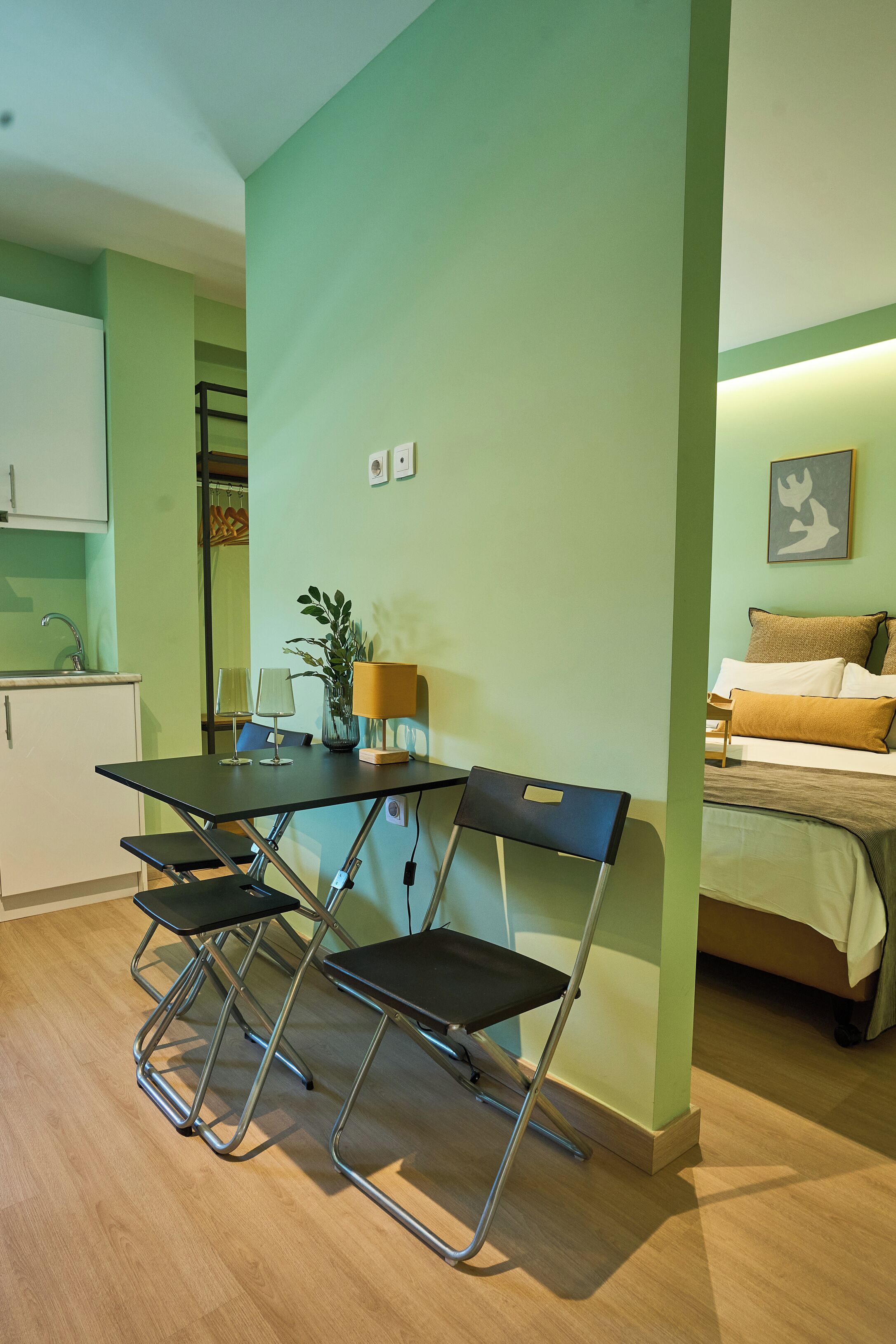 Un espacio habitable compacto con kitchenette, zona de comedor con mesa y sillas, y cama con almohadones.