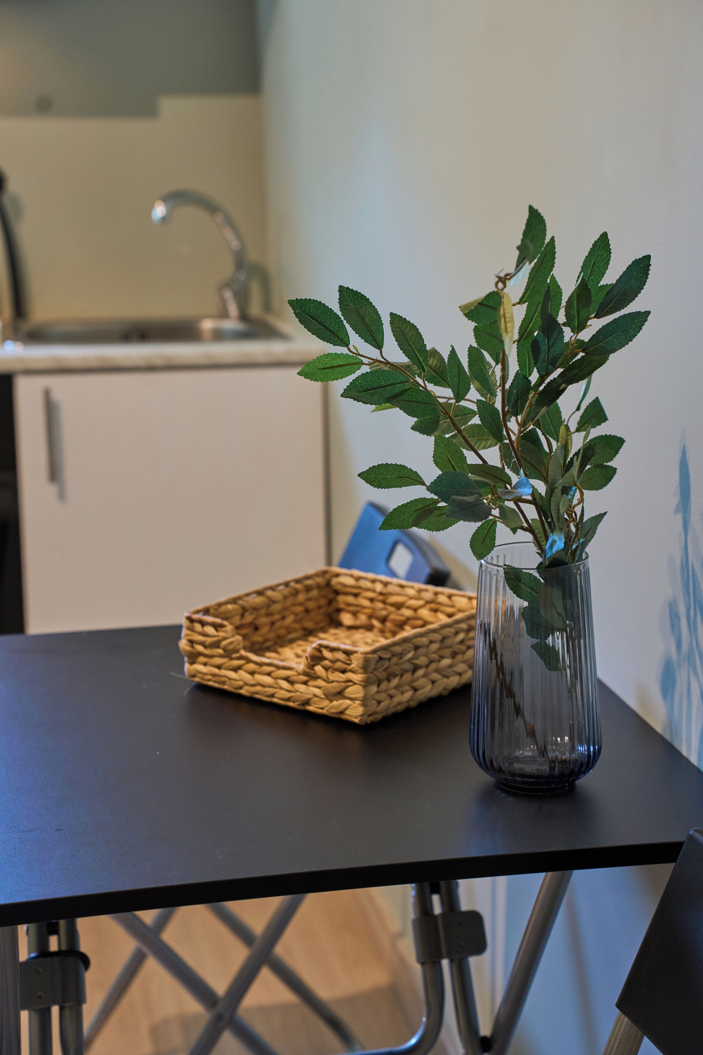 Una cocina con una mesa, una cesta tejida y un florero de vidrio con hojas verdes.