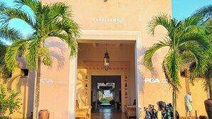 A klubház bejárata, melyet pálmafák szegélyeznek, egy „PGA Akadémia” tábla, és a bejárat előtt golfbag-ek találhatók.