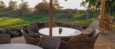 An outdoor seating area with wicker furniture and a table under a large umbrella, overlooking a golf course.