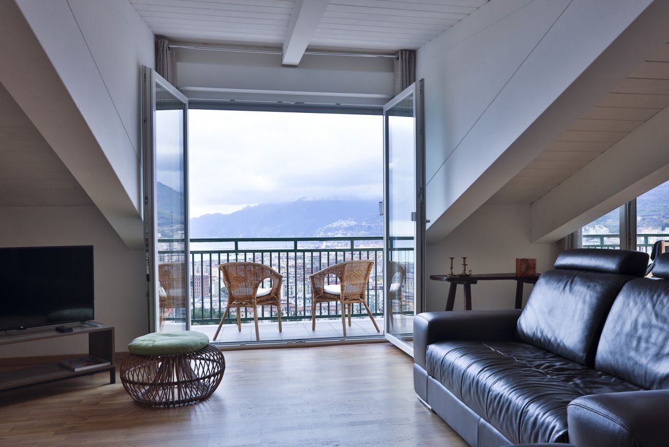 Een moderne woonkamer met een groot raam, een zwarte leren bank, rieten fauteuils en een flatscreen televisie.