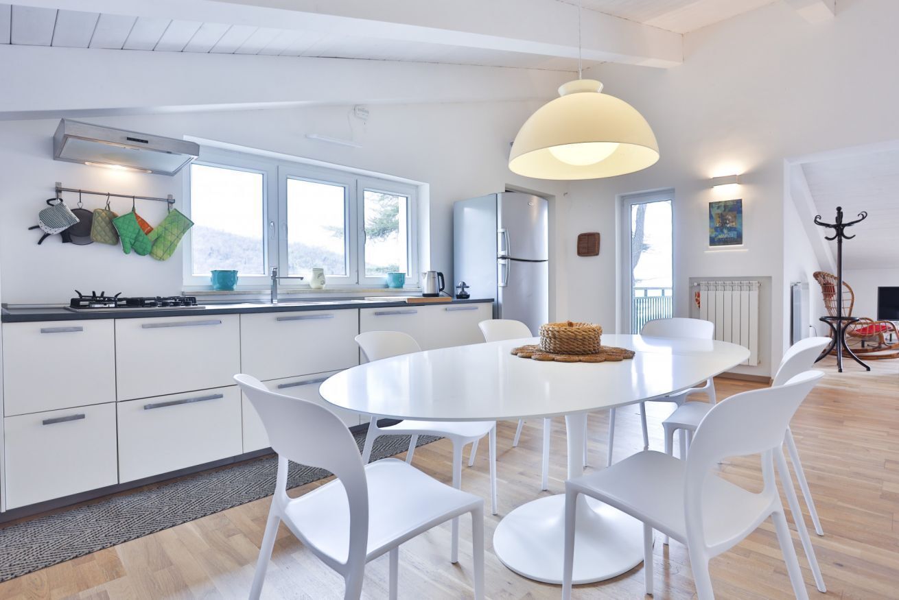 Una cocina moderna con armarios blancos, una mesa de comedor redonda y sillas blancas. Hay un ventanal grande, una lámpara colgante y un perchero.