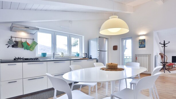 Una cucina moderna con mobili bianchi, un tavolo da pranzo rotondo e sedie bianche. Si nota un'ampia finestra, una lampada a sospensione e un appendiabiti.
