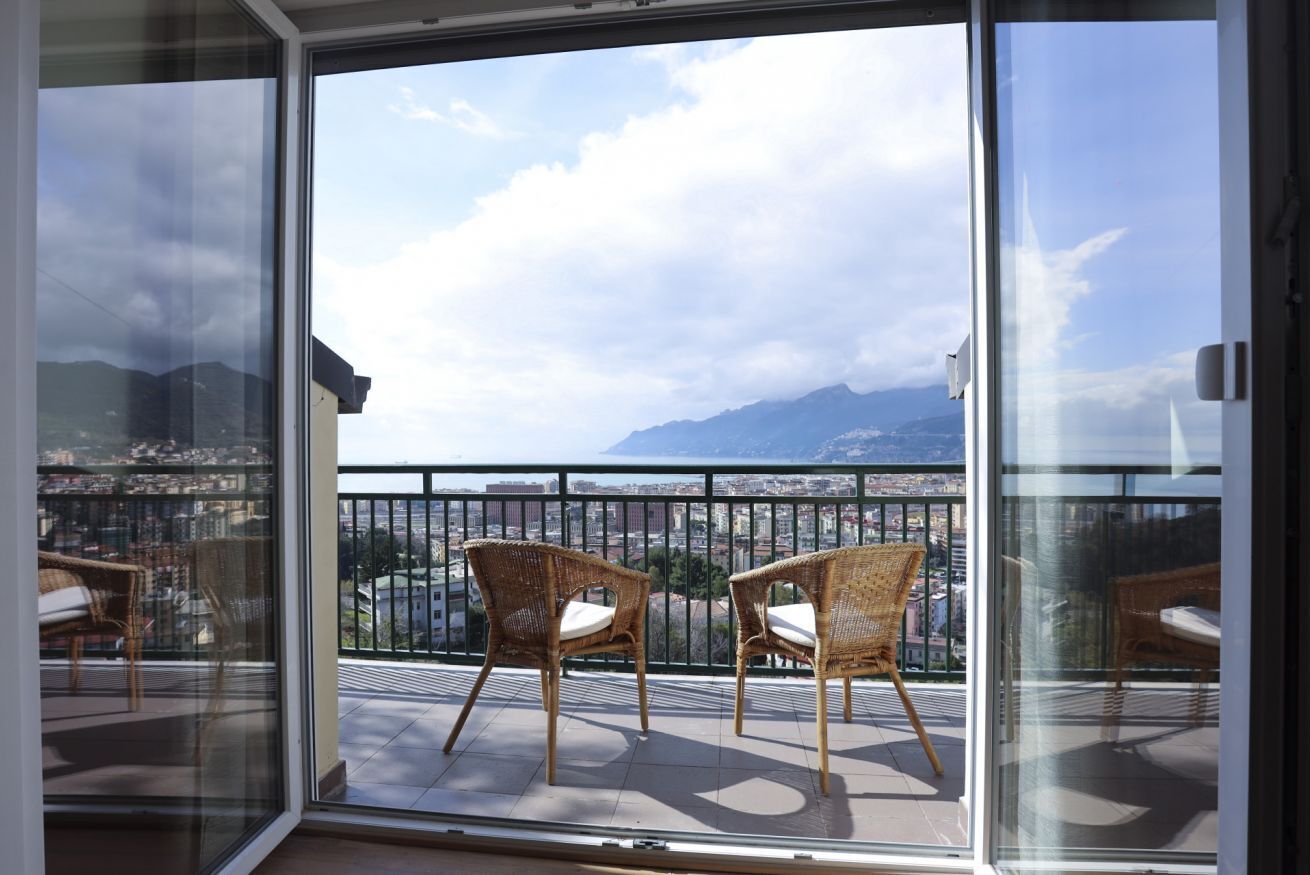 A balcony with wicker chairs, a glass door, and a view of a cityscape and mountains.