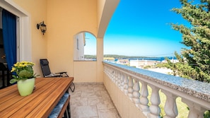 A balcony with a wooden table, a chair, a potted plant, and a view of the sea and buildings. - RS apartment 3 Rab by Villas Guide (Barbat na Rabu)