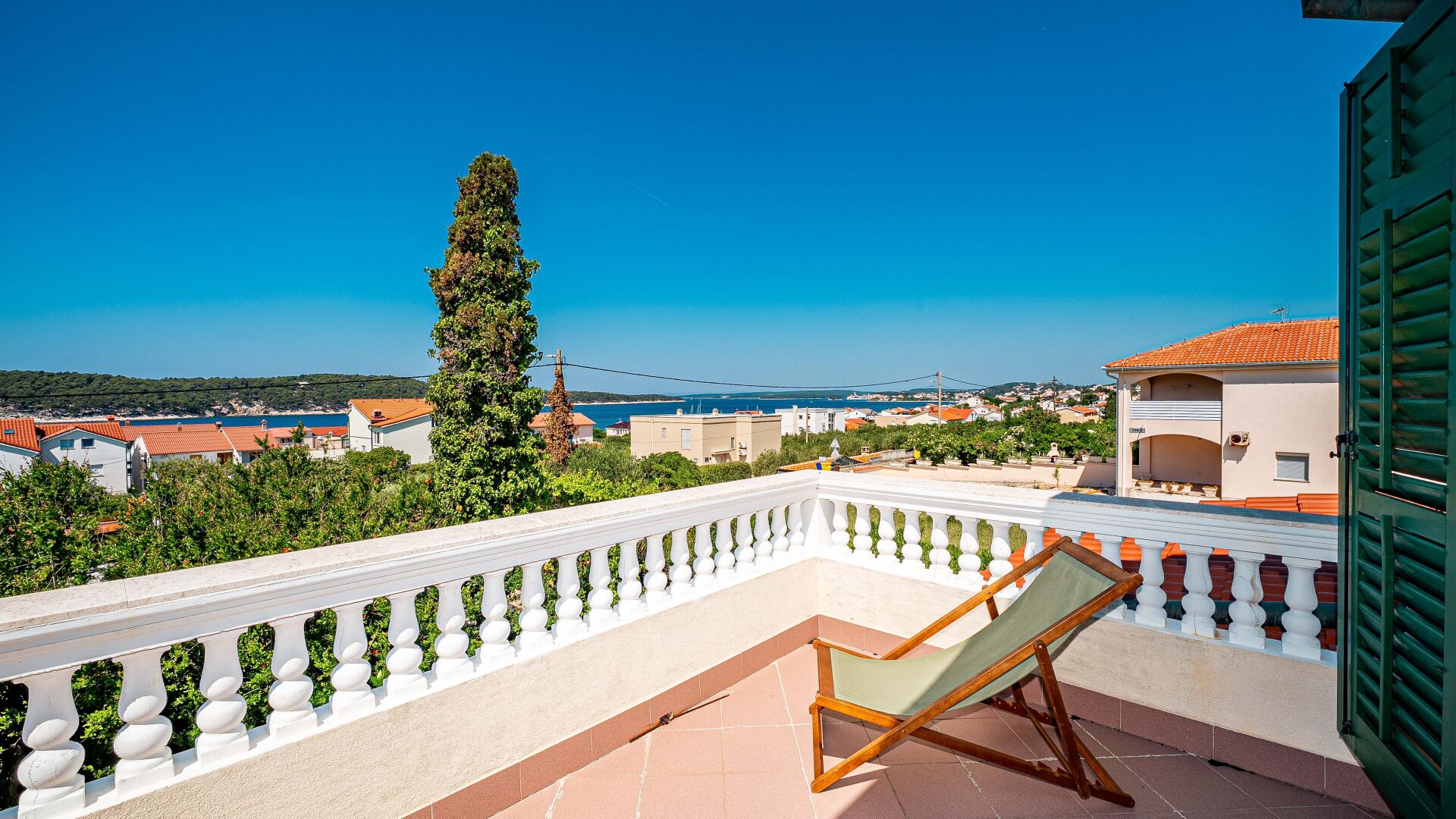 A balcony with a white balustrade, a wooden chair, and a view of a coastal town.
