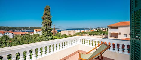 A balcony with a white balustrade, a wooden chair, and a view of a coastal town.