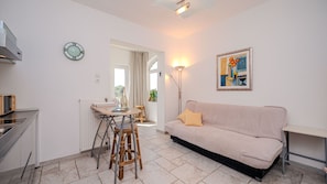 Ein modernes Wohnzimmer mit einem beigefarbenen Sofa, einem Esstisch mit Stühlen, einer Küchenzeile und einem Bild an der Wand.