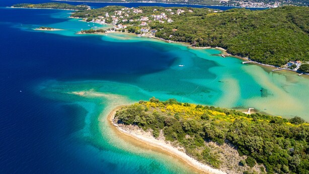 Vista aérea de una zona costera con aguas cristalinas, una playa de arena y una península densamente arbolada.