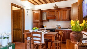 A kitchen with wooden cabinets, a dining table with chairs, a vase with flowers, and a television mounted on the wall.