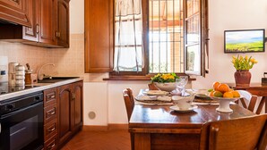 A kitchen with wooden cabinets, a window with curtains, a dining table with chairs, and a bowl of fruit.