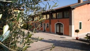 A building with a red facade, a gravel driveway, and a small tree in the foreground. - Corte Belvedere (Sommacampagna)