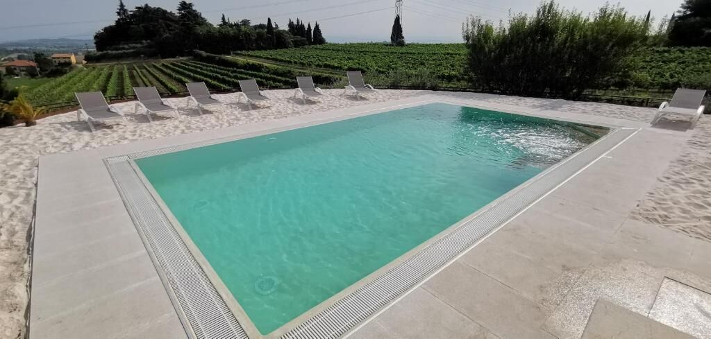 A rectangular swimming pool with clear water, surrounded by white lounge chairs and a sandy area.