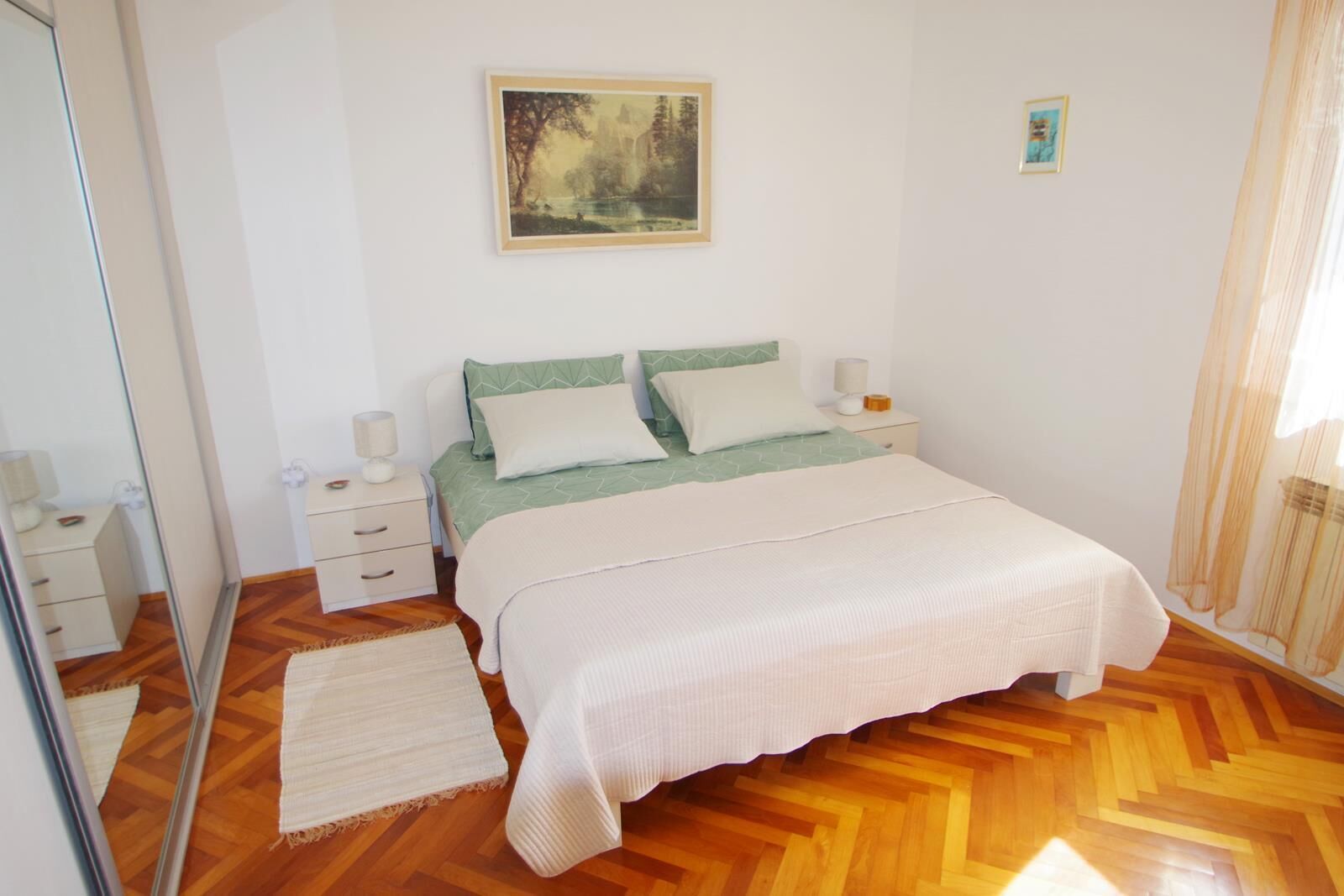 A bedroom with a bed, bedside tables, a mirror, a painting, and a window with curtains.