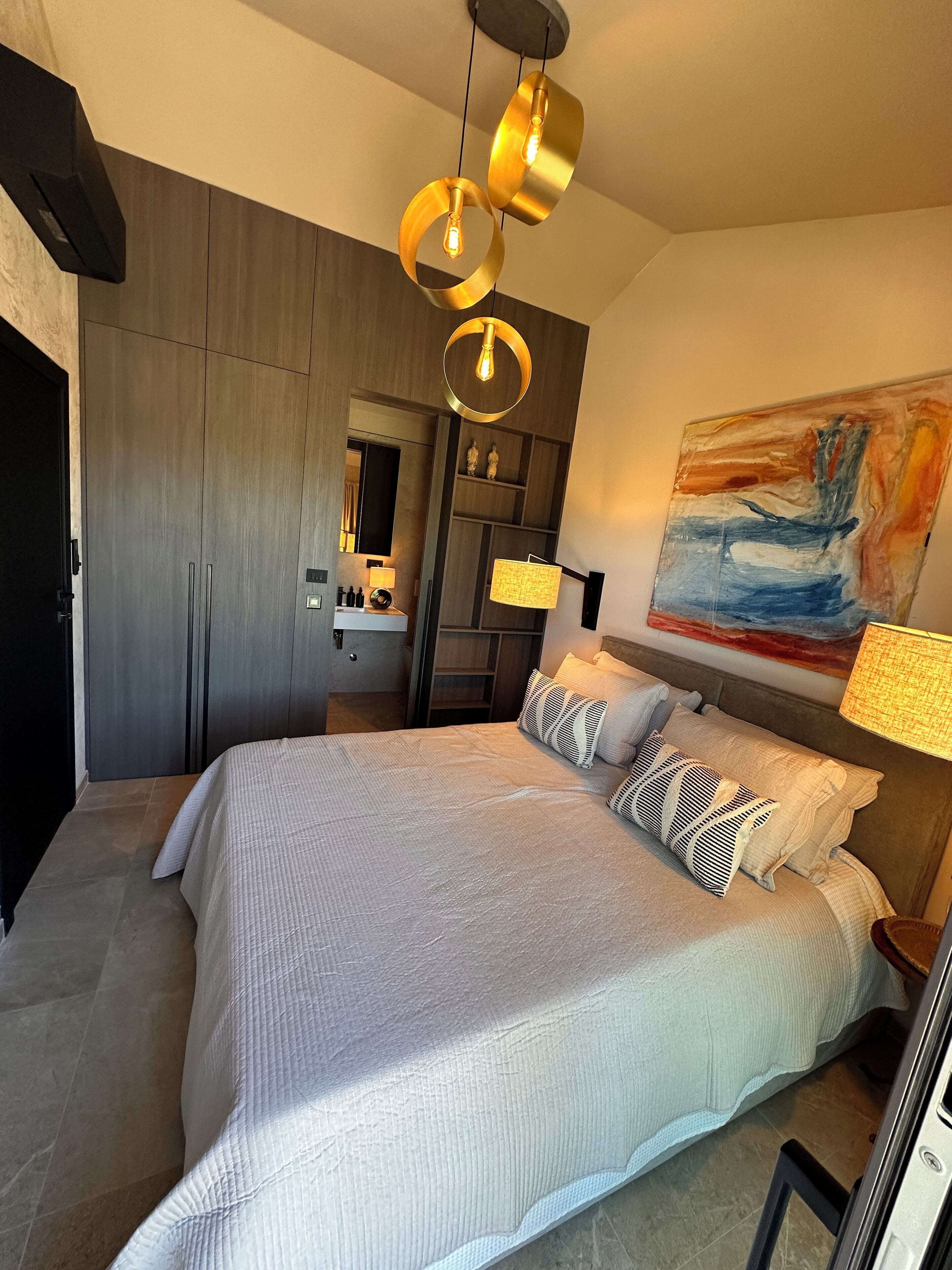 A modern bedroom with a large bed, a wooden wardrobe, a bedside table, a lamp, and a painting on the wall.