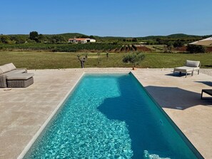 Una piscina rectangular con agua cristalina, rodeada por una solana de hormigón. Hay dos sillones de jardín y un sofá de mimbre en la solana.