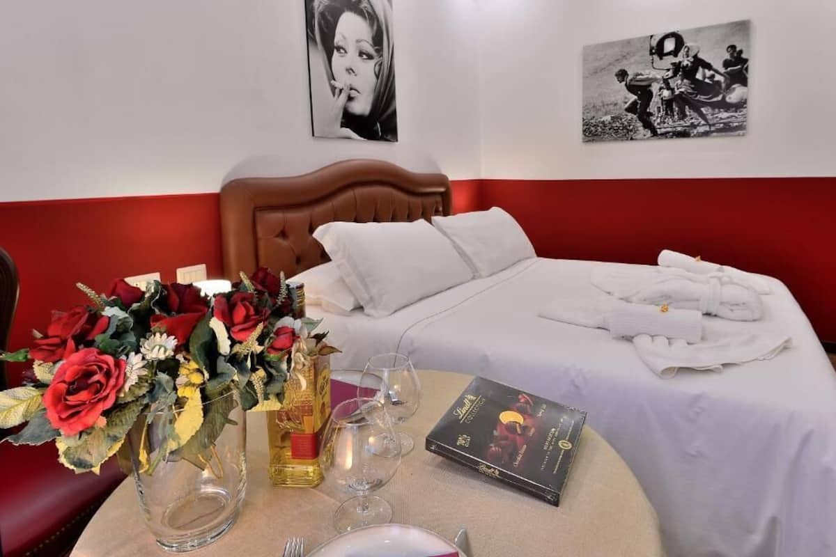 Una cama bien tendida con sábanas blancas, un ramo de rosas rojas, una botella de licor y un libro en la mesita de noche.