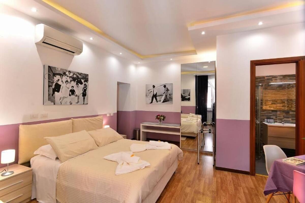 Habitación de hotel con cama, mesita de noche y espejo. Se ve un baño a través de una puerta de cristal.