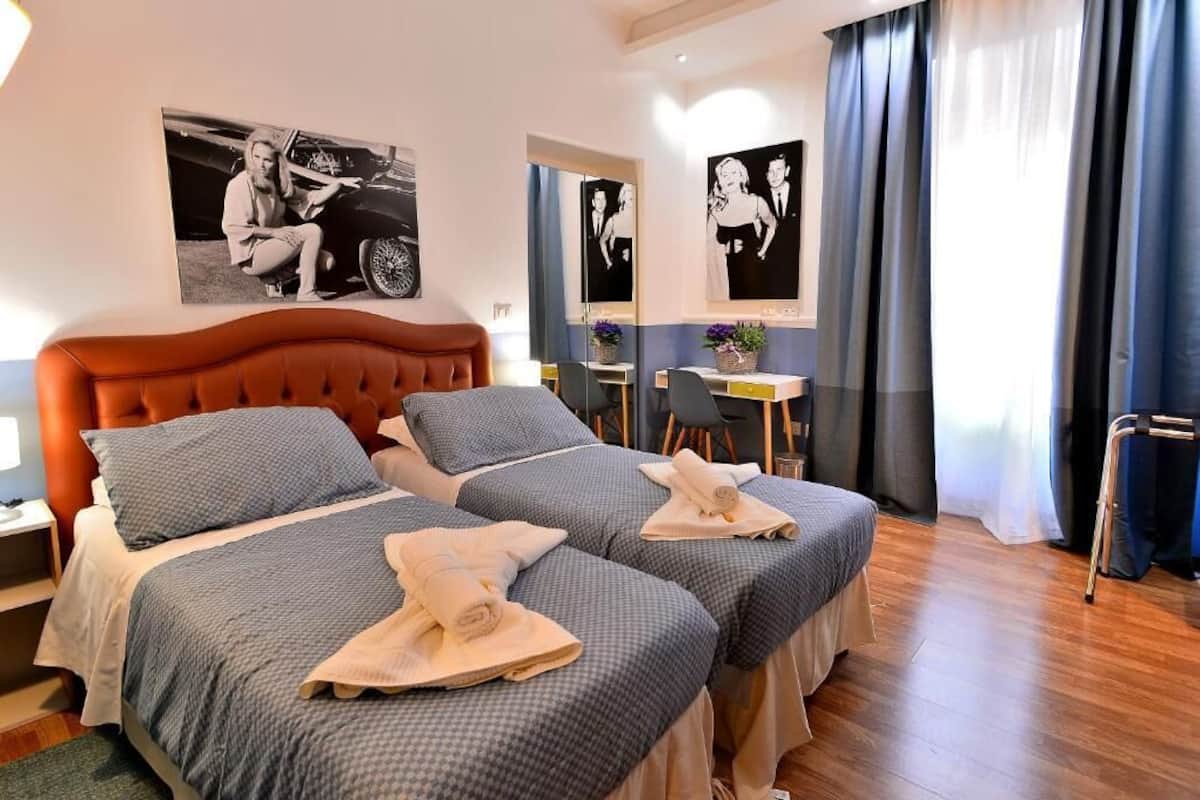 Habitación de hotel con dos camas, un escritorio y una silla. Hay toallas sobre las camas y un cuadro en la pared.