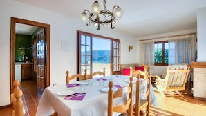 A dining area with a table set for four, wooden chairs, a chandelier, and large windows with a mountain view. - Beautiful House with Stunning Views (Calonge)