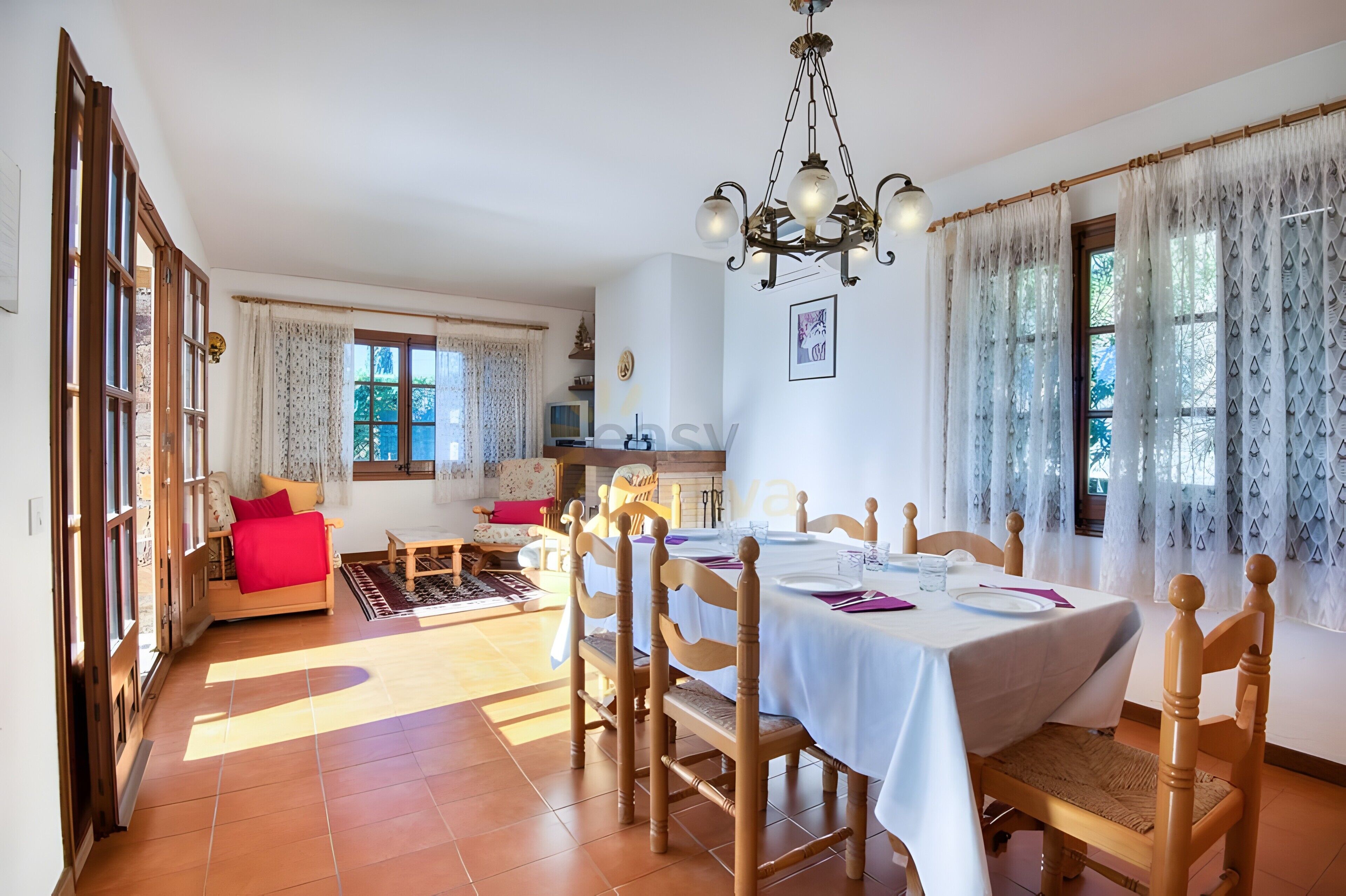 A dining area with a table set for four, wooden chairs, a chandelier, and a view of the garden through the window.