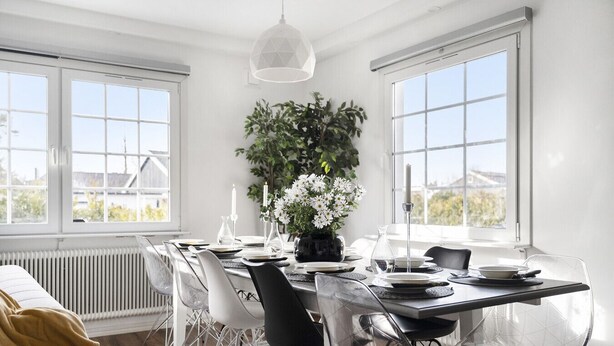 Une salle  à manger moderne avec une grande table dressée pour six personnes, entourée de chaises transparentes. Sur la table, on trouve un pot de fleurs avec des fleurs blanches, et un luminaire suspendu au-dessus.
