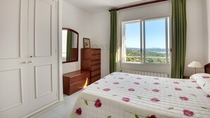 Ein Schlafzimmer mit Bett, Kommode, Spiegel, einem Fenster mit Vorhängen und Blick auf die Landschaft.