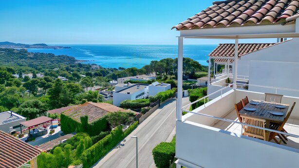 Ein Balkon mit Tisch und Stühlen, der auf eine Küstenlandschaft mit Gebäuden und Grünflächen blickt.