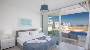 A modern hotel room with a large bed, a bedside table, a view of a swimming pool, and a balcony with a glass door. - 5 & 6 OP Villas Fig Tree Bay Protaras Bedrooms 8, Bathrooms 6, Sleeps 20 (Protaras)