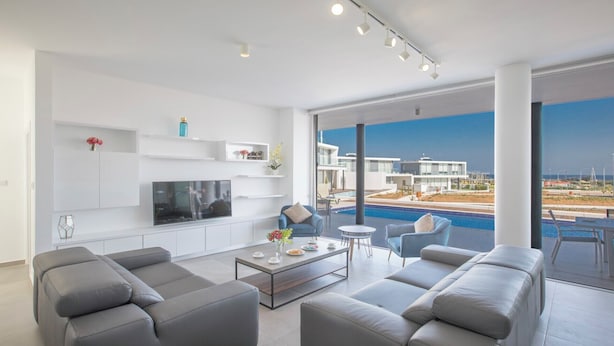 A modern living room with a flat-screen TV, grey sofas, a coffee table, and a pool view. - 5 & 6 OP Villas Fig Tree Bay Protaras Bedrooms 8, Bathrooms 6, Sleeps 20 (Protaras)