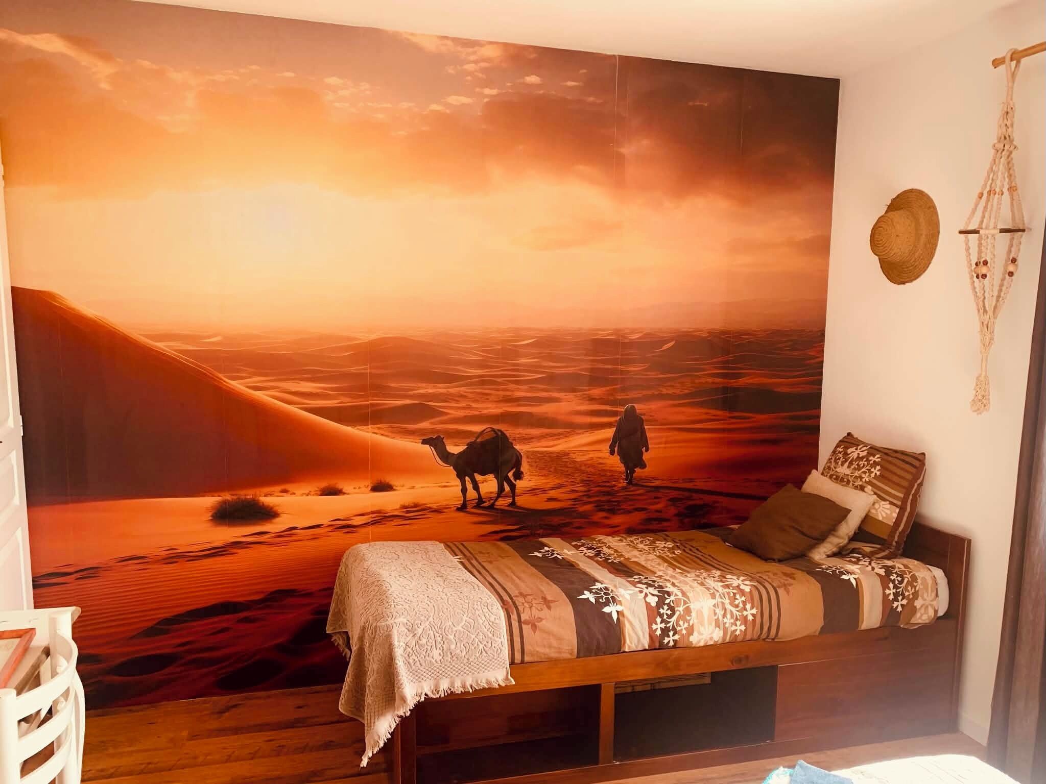 Une chambre avec un grand décor mural représentant un paysage désertique, un lit avec une literie à motifs, une décoration murale en macramé et une table de chevet en bois.