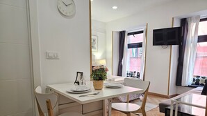 Ein modernes, gut beleuchtetes Zimmer mit einem Essbereich, der einen weißen Tisch, zwei Stühle und einen kleinen Topfpflanzen enthält. Eine Uhr ist an der Wand befestigt und eine TV im Eck sichtbar.